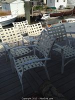 Six white metal frame outdoor chairs with white vinyl webbing seat and backs on a wooden dock by water with boats in background.