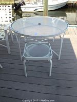 Photo showing round glass topped outdoor table with a white metal frame and a smaller glass topped side table, both on a deck near water.