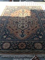 Full view of the rectangular oriental carpet laid out on an outdoor deck. The carpet has brown, blue, and beige floral patterns with a central medallion design and fringes on one end.
