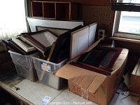 Assorted picture frames in plastic bins and cardboard boxes on a shelf near a window.