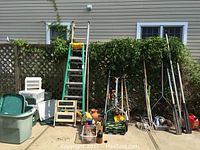Wide shot showing assortment of garden tools, ladders, and plastic bins stored outside against house wall and lattice fence.
