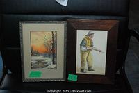 Two framed art pictures on black chair, showing winter landscape on left and man fishing on right.