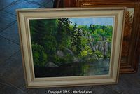 Framed acrylic painting of forest, cliffs, and water on floor.