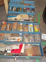 Open blue fishing tackle box with assorted fishing lures, hooks, and tackle showing multiple compartments and trays filled with fishing equipment.