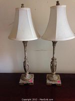 Front view of two matching vintage table lamps showing ornate metal bases and bell-shaped cream fabric shades on dark surface against plain wall.