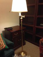 Tall brass floor lamp with a white lampshade standing near wooden furniture and a green couch with a colorful throw.