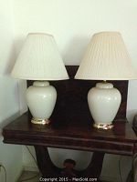 Two white ceramic table lamps with brass bases and beige pleated fabric shades on a wooden table.