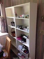 Two white wooden or laminated adjustable shelf bookcases placed side by side, holding various household items on shelves.