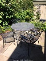 Patio set placed outdoors showing round table with four chairs surrounding it, black wrought iron with mesh design.