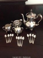 Full tea set displayed on dark wood surface including teapot, two sugar bowls, creamer, sugar tongs, and 12 teaspoons arranged in front.