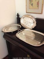 Three silver-toned decorative trays displayed on a dark wood surface: includes oval footed tray with leaf design and inscription, rectangular hammered meat tray, and round tray with detailed edging.