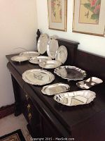 Twelve assorted silver serving trays in various shapes and sizes arranged on a dark wooden sideboard.