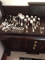 Wide shot of full assortment of silver-tone flatware and serving utensils laid out on a dark wood surface.