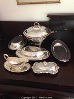 Full view of silver plated set including handle chafing pot, covered dishes, gravy boat, butter dish, and tray on dark surface.