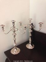 Two silver three-arm candelabras placed on a dark wood surface, showing the overall shape, size, and decorative design.