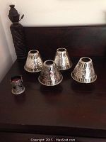 Four silver-tone decorative metal candle shades and one angel-themed metal candleholder with red candle on a dark wooden surface