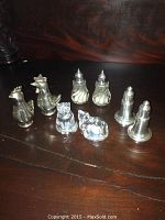 Front view of all five sets of salt and pepper shakers including pewter hens and roosters, glass cats, and pewter birds missing plugs placed on a wooden surface.
