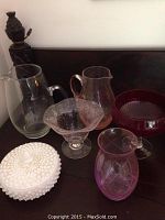 Six assorted glass items grouped: two clear pitchers, hobnail milk glass covered candy dish, clear textured vase or bowl, red glass pedestal bowl, purple etched vase.