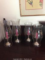 Four hurricane candle holders with pink candles displayed on a wooden surface, two large and two smaller holders.