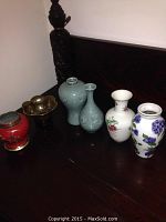 Six Asian decorative vases and bowls displayed on a dark wooden surface, showing the full set and variety in shapes and material.