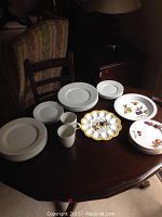 Photo showing the full assortment of white Gibson plates and mugs, colorful Dansk Tapestries patterned plates, Evesham plates with floral motifs, egg holder plate, and one scalloped rooster serving dish on a dark wooden table.