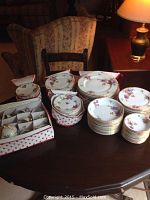 Porcelain china set arranged on a wooden table including cups in a box, stacked plates, saucers, and bowls with floral designs and gold trim.