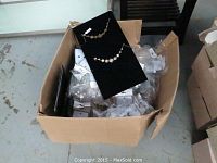 Wide view of box containing numerous sealed packs of jewelry and two floral design gold-tone bracelets displayed on black velvet boards.
