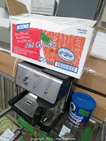 Krups coffee maker with glass carafe on a metal drip tray, box of Dixie hot cup lids, and Maxwell House coffee canister nearby.