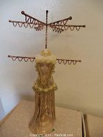 Photograph showing a mannequin-shaped jewelry holder with metal arms having multiple hooks for hanging jewelry items. The mannequin is decorated with lace and fringe.