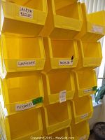 Stacked yellow plastic storage bins with various handwritten labels on the front of each bin.