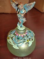 Close-up of a greenish glass perfume bottle decorated with blue flower patterns and colorful gem accents, topped with a detailed winged cherub figurine.