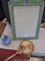 Green decorative picture frame, vintage style hand mirror, and blue flower-themed perfume bottle visible on table.