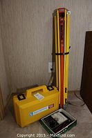 Collapsed yellow and red metal tripod standing next to yellow plastic carrying case and multimeter box against wall.