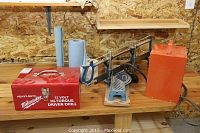 Wide shot of all three items on a wooden bench including the red metal heater, blue Mastercraft mitre saw, and red Milwaukee toolbox case.
