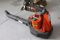 View of orange Husqvarna gas leaf blower with attached black blower tube on a concrete floor showing the blower from side angle.