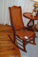 Side view of antique nursing rocker showing wooden frame, caned seat and backrest, and curved rockers on hardwood floor near a side table.