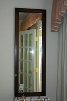 Full view of the wood framed mirror hanging vertically on the wall, showing reflection of a door and window with a curtain.