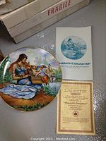 Photo of Pharaoh's Daughter plate along with certificate and original box showing detail artwork of woman beside a river holding baby Moses.