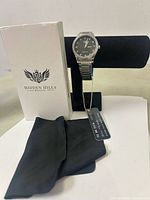 Photo of the watch displayed on a black stand, accompanied by the white branded box and black cleaning cloth.