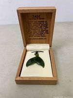 Dark green jade whale tail pendant necklace displayed in an open wooden box with ivory interior and engraved informational text about jade.