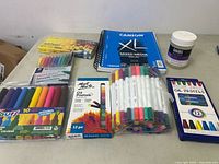 Full lot showing all art supplies spread out on table including pastel markers, oil pastels, mixed media sketchbook, and white container of paste medium.