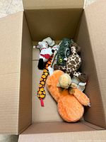 Cardboard box open showing a collection of various stuffed animals including orange monkey, giraffe, dog, horse, plush snake and a green alligator toy keychain