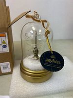 Clear glass dome ornament with gold base and attached Harry Potter tag and hanging ribbon shown on white foam placement.