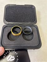 Open black Ridge branded protective case containing two rings: one 24 karat gold plated beveled ring and one black ring, both new and unused.