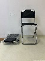 Two new mini folding chairs, one unfolded and standing, one folded and wrapped in plastic, against plain wall.