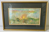 Framed abstract watercolor landscape painting with a dark green mat and gold frame showing a sunset behind hills and some small structures below.