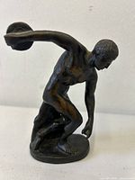 Front left side view of bronze statuette of a nude discus thrower in action pose on oval base.