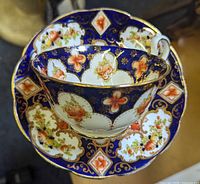 Top view of teacup placed on saucer showing the floral and geometric Imari style pattern with gold accents and dark blue background.