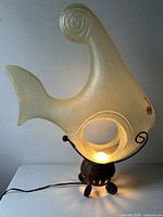 Front view of the illuminated acrylic angel fish table lamp on the metal base, showing the curled tail and warm glow from the light source inside the acrylic body.