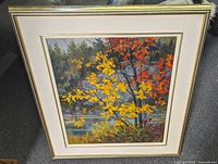 Framed oil painting showing vibrant autumn trees by a shoreline with yellow and orange leaves. Frame is off-white with gold trim.
